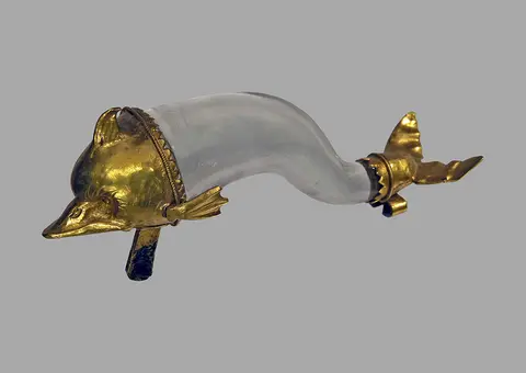 A decorative vessel shaped like a dolphin, with a transparent body and gold accents. The dolphin's head and tail are elaborately designed, showcasing intricate detailing in the gold material. The overall appearance combines elegance and craftsmanship in this unique art piece.