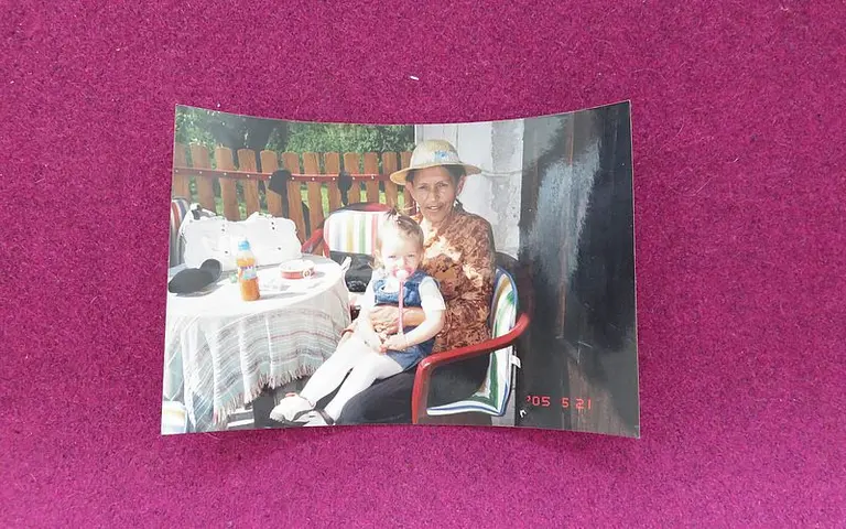 Ein älterer Mensch sitzt auf einem Stuhl neben einem kleinen Mädchen. Sie teilen einen Tisch mit Getränken und einem weiteren Gegenstand. Der Hintergrund zeigt einen Garten mit einem Holzzaun. Beide wirken entspannt und glücklich.