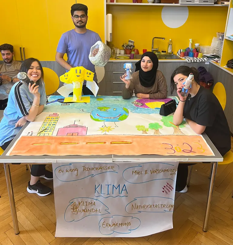 Eine Gruppe von vier Personen steht um einen Tisch, auf dem ein buntes Plakat über Klima und Umweltprobleme präsentiert wird. Sie halten verschiedene Gegenstände in der Hand und zeigen auf das Plakat, das Themen wie Wassermangel und Umweltverschmutzung behandelt.