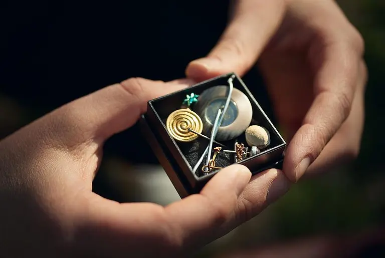Eine Person hält eine kleine, offene Box in den Händen, die verschiedene dekorative Objekte enthält, darunter eine spiralartige goldene Figur, ein blauer Stein, einen kleinen Gegenstand und eine Nadel. Die Box ist auf einem dunklen Untergrund platziert.