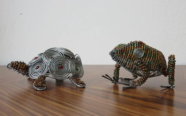 Zwei kunstvoll gestaltete Frosch-Skulpturen aus Draht stehen auf einer Holztischoberfläche. Der linke Frosch ist silberfarben mit spiralförmigen Mustern, während der rechte Frosch in verschiedenen Farben mit bead-artigen Details verziert ist.