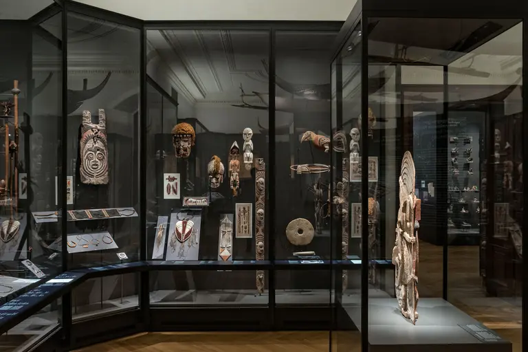 Im Bild sind mehrere Vitrinen in einem Museum zu sehen, die eine Vielzahl von ethnographischen Artefakten ausstellen. Die Objekte umfassen Masken und Statuen, die künstlerisch gestaltet sind und verschiedene kulturelle Elemente repräsentieren. Der Raum hat eine ruhige Atmosphäre, die die Exponate hervorhebt.