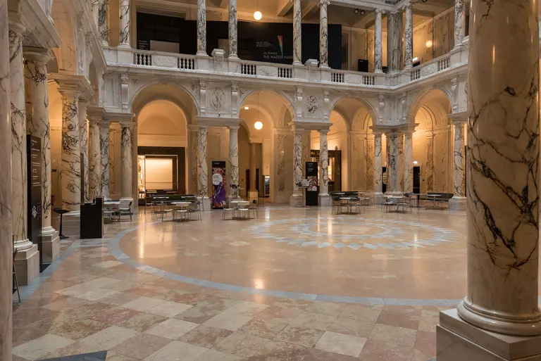 Ein weitläufiger Innenraum mit hohen Säulen aus Marmor und einem dekorativen Boden. In der Mitte des Raumes ist ein runder Bereich mit künstlerischen Motiven. Die Wände sind mit Gemälden geschmückt, und Tischgruppen sind in der Mitte angeordnet.