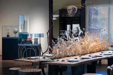 Ein modern gestalteter Raum mit einem langen Holztisch, der mit Tellern gedeckt ist. In der Mitte befindet sich eine dekorative Anordnung aus getrocknetem Gras und Pflanzen. Im Hintergrund stehen Glasvitrinen mit Ausstellungen und einem weiteren Tisch mit einer Skulptur.
