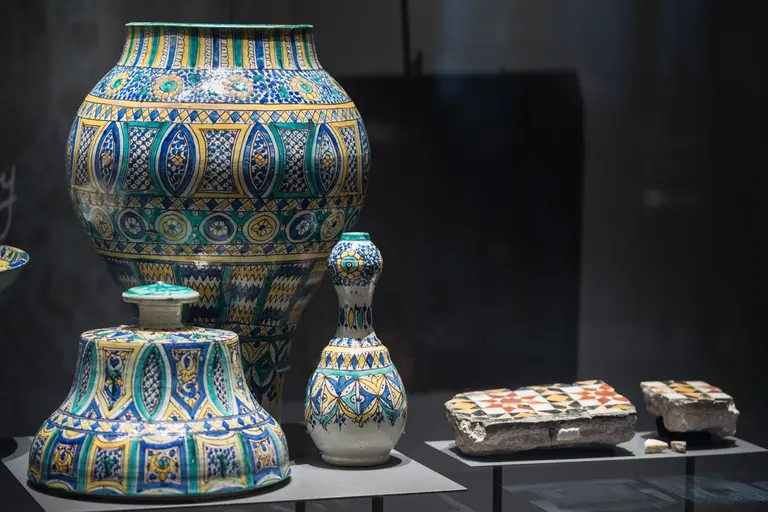 In der Abbildung sind verschiedene kunstvoll verzierte Keramiken zu sehen. Dazu gehören bunte Vasen mit geometrischen Mustern und eine schmalere Flasche. Die Objekte zeigen detaillierte Handwerkskunst und sind auf einem Ausstelltisch zu sehen.