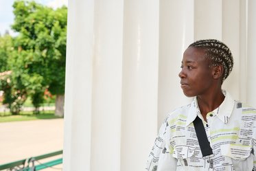 Eine Person mit kurzen, geflochtenen Haaren steht neben einer hellen Säule und schaut nachdenklich in die Ferne. Im Hintergrund sind grüne Bäume zu sehen, und der Himmel ist klar.