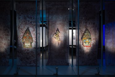 Three illuminated tapestries are displayed behind glass panels, featuring intricate designs. The tapestries are in the shape of pointed arches, showcasing colorful and detailed illustrations against a textured, marbled background in a dimly lit setting.