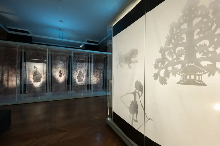 An art exhibit showcases illuminated shadow puppets and intricate silhouettes against textured backgrounds. The display features various characters and scenes, highlighting the unique interplay of light and shadow in a creative setting. The atmosphere combines artistry with cultural storytelling.
