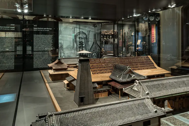 In der Ausstellung sind Modelle historischer Gebäude zu sehen, die Einblicke in die Architektur vergangener Zeiten geben. Im Hintergrund sind informative Texte und Illustrationen zu erkennen, die die Geschichte der dargestellten Objekte erläutern.