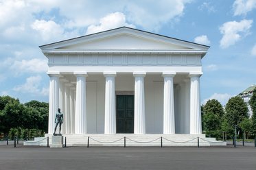 Ein klassisches Gebäude mit weißen Säulen und einem schlichten Dach steht unter einem blauen Himmel. Vor dem Gebäude befindet sich eine Statue. Der Boden ist asphaltiert und es gibt eine Absperrung um das Gebäude herum.