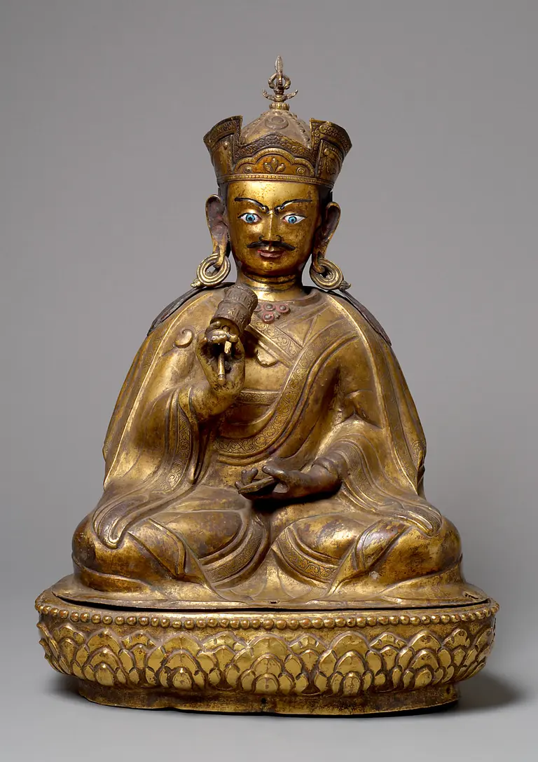 Eine goldene Statue eines sitzenden buddhistischen Meisters mit einem königlichen Kopfschmuck. Er hält ein rotes Objekt in der linken Hand und hat detailreiche Gesichtszüge, darunter blaue Augen. Die Statue steht auf einem kunstvoll verzierten Sockel.