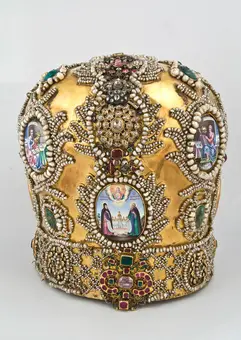 A richly decorated golden crown adorned with intricate patterns, pearls, and colorful gemstones. The crown features several small images encased in ornate frames, depicting religious figures and scenes. Its elaborate design highlights craftsmanship and royal significance.