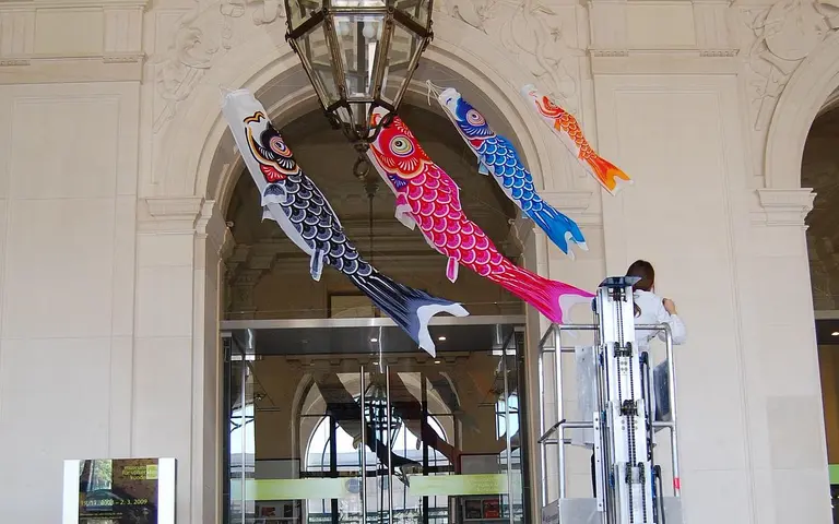 A person on a ladder installs colorful koi fish banners hanging from an archway inside a building. The banners are predominantly pink, red, blue, and black, arranged in a vertical line. The setting features a large chandelier and architectural details typical of indoor spaces.
