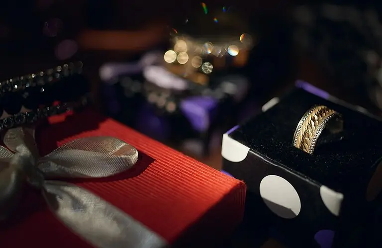 Ein setzendes Bild mit mehreren Geschenken in verschiedenen Farben. Im Vordergrund ist ein roges Paket mit einer Schleife zu sehen. Außerdem liegt ein goldener Ring auf einem schwarzen Samtbezug, umgeben von verschwommenen dekorativen Elementen.