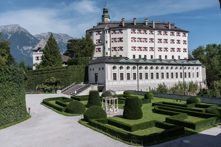 Ein historisches Gebäude mit mehreren Stockwerken und grünen Fensterläden steht vor einem klaren Himmel. Im Vordergrund befinden sich kunstvoll gestaltete Gartenanlagen mit geometrischen Hecken und einem Pavillon. Im Hintergrund sind Berggipfel sichtbar, die eine beeindruckende Kulisse bieten.