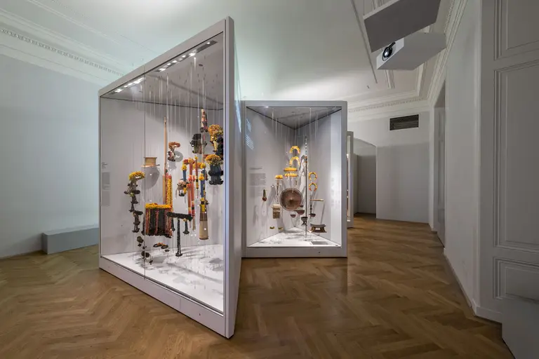 Zwei gläserne Vitrinen in einem Ausstellungsraum zeigen kunstvolle Objekte. Die Vitrinen enthalten verschiedene Gegenstände, die vielfältige Formen und Farben aufweisen. Der Boden besteht aus Holz, und die Wände des Raumes sind schlicht und hell.