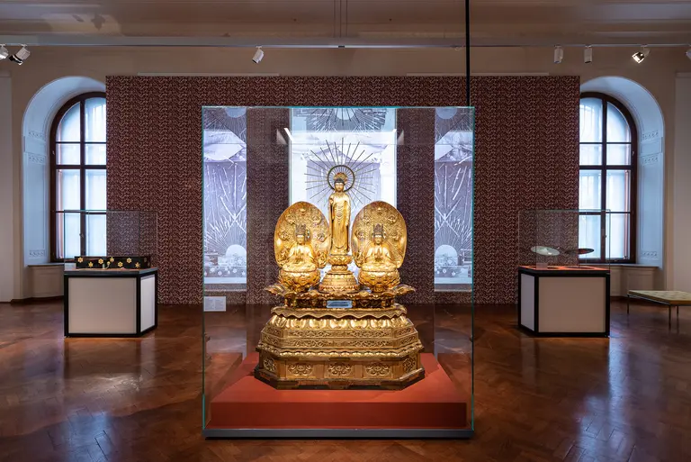 A golden statue of a seated figure with a halo is prominently displayed in a glass case. Flanking the statue are two smaller figures. The background features decorative walls, and additional objects are shown in nearby display cases within a museum setting.