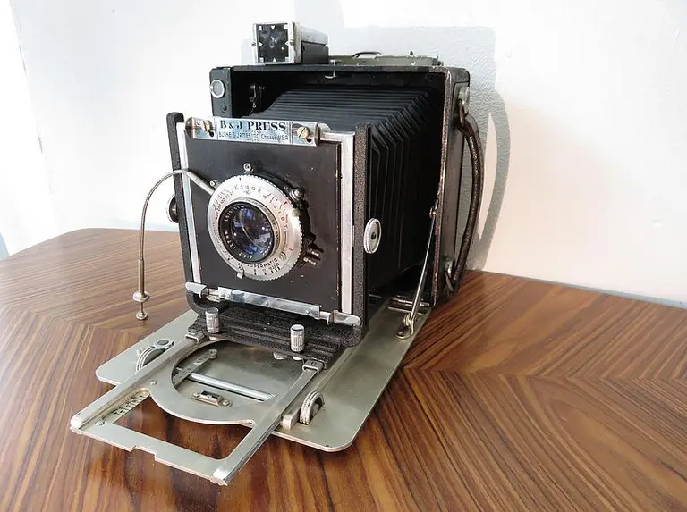 Eine klassische Großformatkamera steht auf einem Tisch. Sie hat einen großen quadratischen Körper mit einem ausgeklappten Objektiv und einer Faltmechanik. Die Kamera ist schwarz und silber, und das Objektiv ist gut sichtbar in der Mitte platziert.