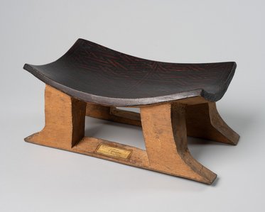 Ein traditioneller Holzsitz mit einer gewölbten Oberfläche und zwei stabilen Beinen. Die Sitzfläche ist schwarz mit roter Verzierung, während die Beine aus hellem Holz bestehen. Ein kleines Namensschild ist an der Vorderseite angebracht.