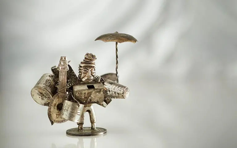 Eine kleine Skulptur aus Metall zeigt einen Mann, der mit einer Vielzahl von Werkzeugen und Geräten beladen ist. Er trägt einen Hut und hält einen Regenschirm in der Hand. Die Figur steht auf einer glatten Oberfläche vor einem hellen Hintergrund.