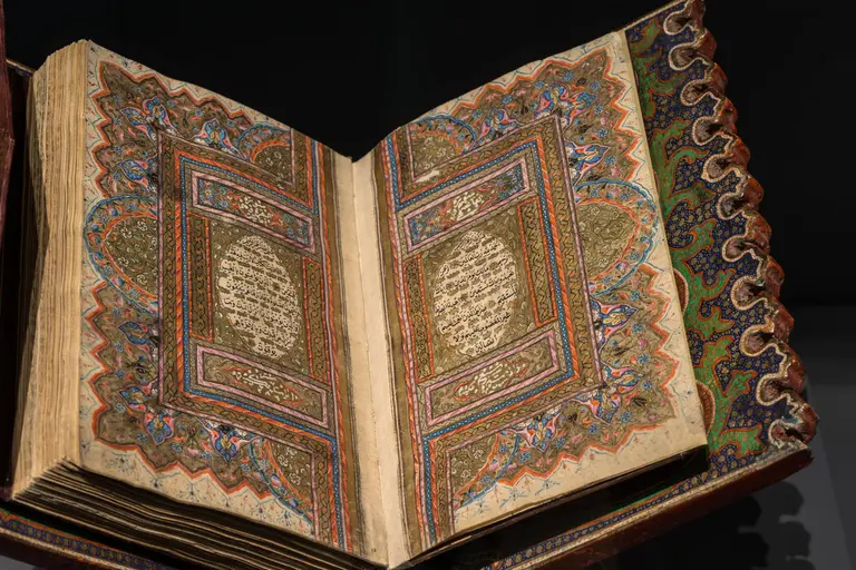 Das Bild zeigt ein geöffnetes, kunstvoll verziertes Buch mit detaillierten, farbenfrohen Illustrationen. Die Seiten sind mit arabischem Text und ornamentalen Mustern geschmückt, die eine traditionelle und historische Handschrift widerspiegeln.