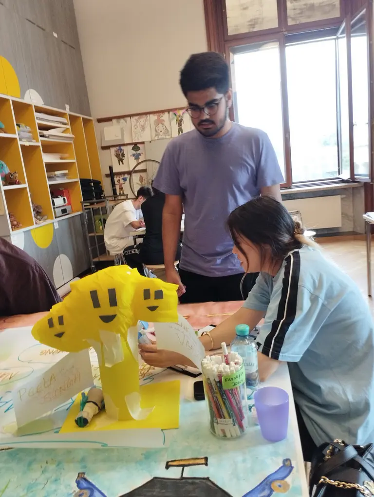 Zwei Personen arbeiten an einem Bastelprojekt. Eine Person sitzt am Tisch und malt an einer gelben Figur, die wie ein Smiley-Baum aussieht. Die andere Person steht daneben und beobachtet. Im Hintergrund sind weitere Aktivitäten zu sehen, aber der Fokus liegt auf den beiden Bastlern.