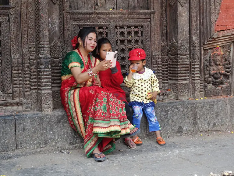 Eine Frau in traditioneller Kleidung sitzt auf einer Steinmauer und schaut mit zwei Kindern auf ein Handy. Beide Kinder sind spielerisch und die Umgebung hat kunstvolle Holzschnitzereien. Die Stimmung ist freundlich und familiär.
