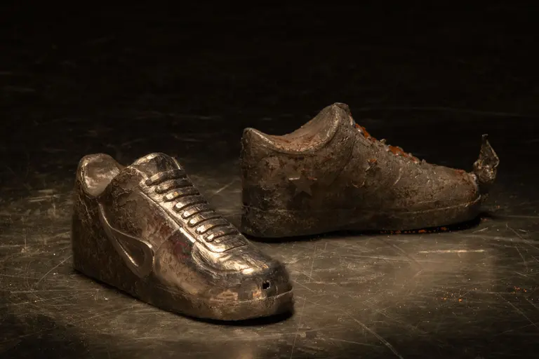 Zwei Schuhe aus Metall stehen auf einem dunklen Boden. Der linke Schuh hat eine glatte, glänzende Oberfläche, während der rechte Schuh stark abgenutzt und rostig aussieht. Beide Schuhe sind stilisiert und nicht tragbar.
