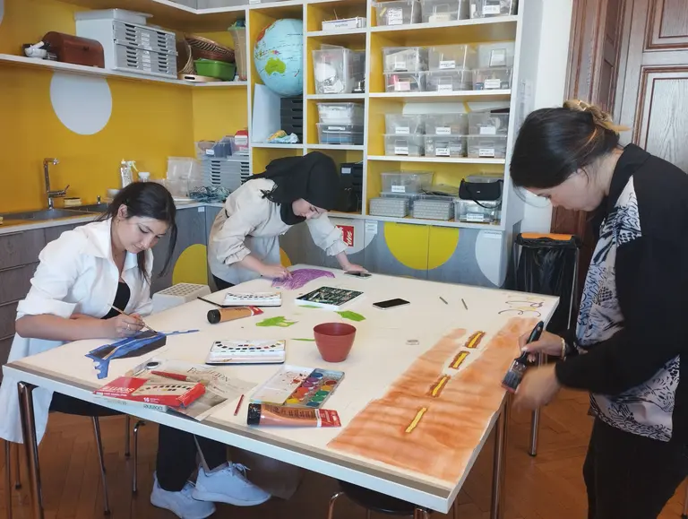 Drei Frauen sitzen an einem Tisch und arbeiten kreativ. Eine malt mit Wasserfarben, während die andere mit Stiften zeichnet. Die dritte Frau nutzt ein Werkzeug, um an ihrem Projekt zu arbeiten. Auf dem Tisch sind verschiedene Materialien und Farben verteilt. Die Umgebung wirkt freundlich und einladend.