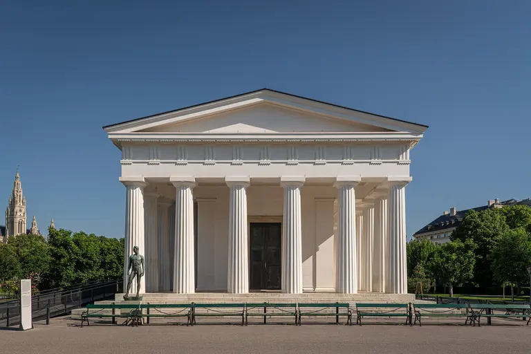 Ein neoklassizistisches Gebäude mit hohen, weißen Säulen und einer breiten Treppe. Im Vordergrund stehen Bänke und links ist eine Statue zu sehen. Im Hintergrund sind Bäume und ein weiterer Gebäudeteil zu erkennen. Der Himmel ist klar und blau.