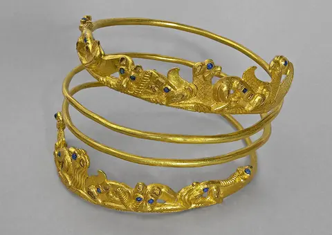 Three intertwined gold bracelets featuring ornate designs with small blue stones embedded throughout. The bracelets are crafted to resemble natural elements like waves or foliage, showcasing intricate detailing and a shiny finish. The background is a soft gray, enhancing the jewelry's lavish appearance.