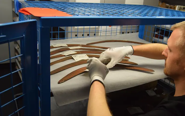 A person wearing gloves carefully handles wooden objects inside a blue storage crate. The items are laid out on white paper, with labels attached. The setting appears to be a museum or archival storage area, focused on preserving artifacts.