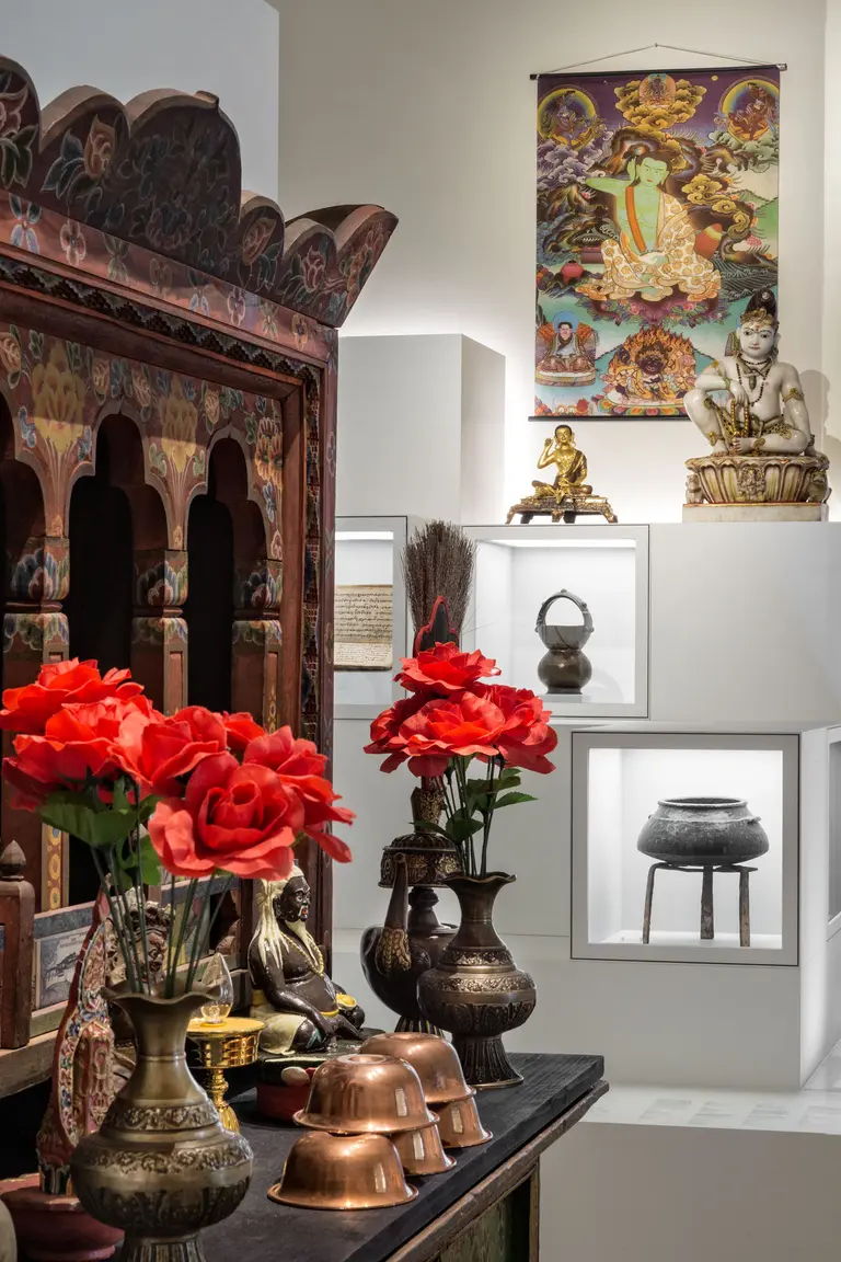 Eine Komposition aus traditionellen Artefakten und Kunstwerken, darunter eine verziertes Holzregal, Kupfergefäße, und eine Statue. Rote Blumen schmücken den Tisch, während ein Wandbild und weitere Statuen im Hintergrund zu sehen sind.