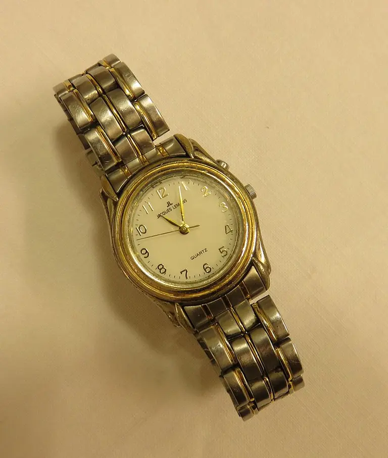 Eine elegante Armbanduhr mit einem runden, goldenen Gehäuse und einem hellen Zifferblatt. Die Ziffern sind schwarz, und die Uhr hat einen goldenen Sekundenzeiger. Das Armband besteht aus einem Metall in Edelstahloptik, das in Gliederform gestaltet ist.