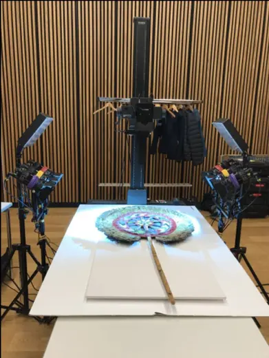 Das Bild zeigt einen Tisch mit einer großen, bunten Kreisspirale und einem langen Stock in der Mitte. Über dem Tisch sind Kameras und Lichtquellen positioniert, die auf das Objekt ausgerichtet sind. Im Hintergrund sind Holzpaneele zu sehen.