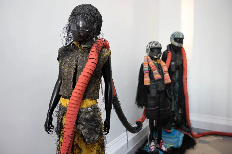 Drei Schaufensterpuppen stehen in einem hellen Raum. Die Puppen sind kreativ mit unterschiedlichen Textilien und farbigen Schläuchen gestaltet. Die erste Puppe trägt ein schwarzes Oberteil mit gelbem Rock, während die anderen Puppen modische Kopfbedeckungen und farbenfrohe Schals haben.