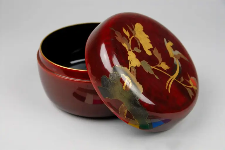 A decorative red lidded bowl with intricate gold floral designs on the lid. The bowl is shown partially open, revealing a smooth black interior. The glossy finish highlights the craftsmanship and elegance of the piece.