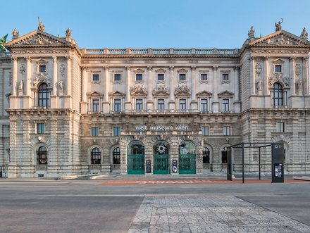 The image shows the ornate facade of a historic building, known as the Weltmuseum Wien, in Vienna. The architecture features intricate decorations, large arched windows, and a grand entrance. The sky is clear, highlighting the details of the structure against a serene backdrop.