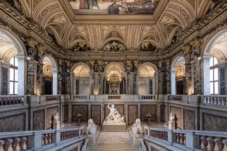 Die Aufnahme zeigt die prächtige Eingangshalle eines historischen Gebäudes mit hohen Decken, großen Fenstern und kunstvollen Verzierungen. Im Mittelpunkt steht eine monumentale Skulptur, umgeben von eleganten Säulen und einer einzigartigen Architektur, die den gesamten Raum ausfüllt.