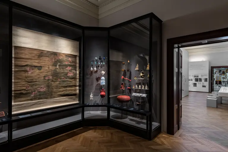 In einem Ausstellungsraum sind Vitrinen mit verschiedenen Objekten zu sehen, darunter ein großes Wandteppich und kunstvolle Artefakte. Der Raum hat einen eleganten Holzfußboden und zeigt weitere Exponate in der Hintergrundansicht.