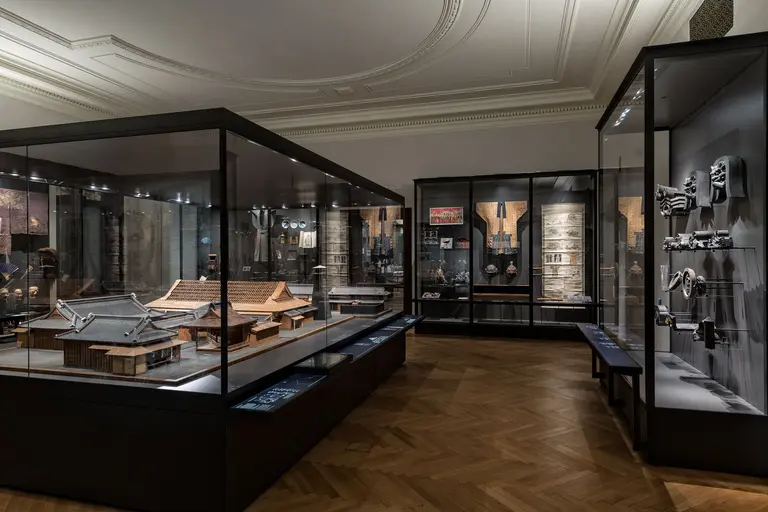 Das Bild zeigt den Innenbereich eines Museums mit Vitrinen, die historische Modelle und Artefakte ausstellen. Im Vordergrund sind Holzmodelle von Gebäuden zu sehen, während im Hintergrund verschiedene Objekte an Wänden präsentiert werden. Die Ausleuchtung hebt die Exponate hervor.