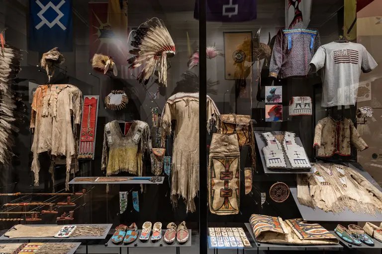 Eine Ausstellung mit traditionellen Kleidungsstücken, Kunstwerken und handgefertigten Objekten amerikanischer Ureinwohner. Zu sehen sind Bemalungen, Federschmuck und Kleidung aus verschiedenen Materialien, die die kulturelle Vielfalt und das Erbe dieser Gemeinschaften repräsentieren.
