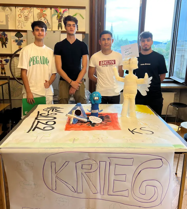 Vier Jugendliche stehen an einem Tisch. Der Tisch ist mit einem großen Banner beschriftet, auf dem das Wort