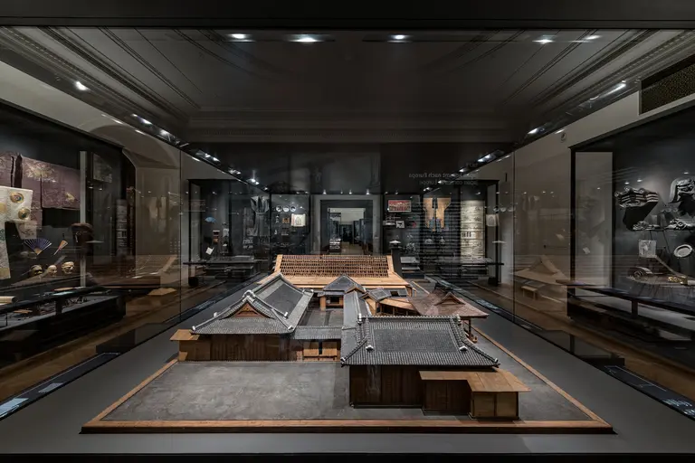 Das Bild zeigt eine Vitrine in einem Museum mit einem detaillierten Modell eines traditionellen japanischen Holzhauses. Im Hintergrund sind weitere Exponate und Wände sichtbar, die verschiedene kulturelle Artefakte präsentieren. Die Beleuchtung hebt die Struktur des Modells hervor.