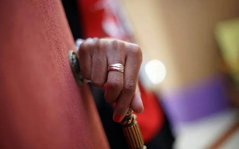 Eine Hand mit einem Ring greift nach einem Türgriff. Der Hintergrund ist unscharf und zeigt verschiedene Farben, die näheren Details sind nicht sichtbar.