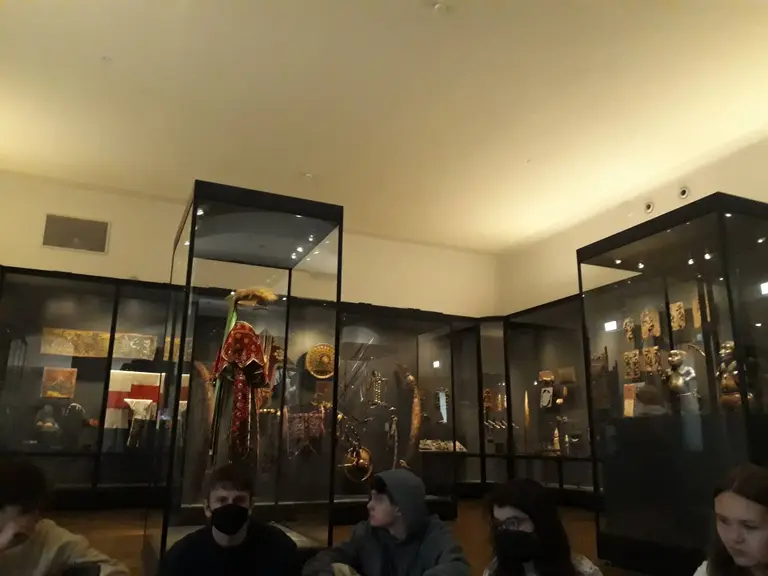 In einem Museum sind mehrere Vitrinen mit historischen Exponaten zu sehen. Im Vordergrund sitzen Besucher auf dem Boden, während sie die Ausstellungen betrachten. Die Vitrinen enthalten verschiedene Objekte und Kunstwerke, die hinter Glas präsentiert werden.