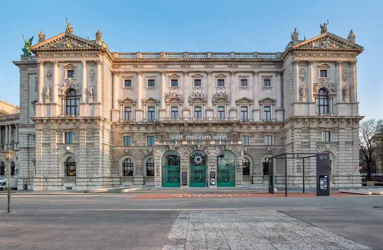 Das Bild zeigt die Fassade des Weltmuseums Wien. Das historische Gebäude ist reich verziert und hat große Fenster mit grünem Rahmen. Über dem Eingang prangt der Schriftzug „welt museum wien“. Der Himmel ist klar und blau, was eine helle Atmosphäre schafft.