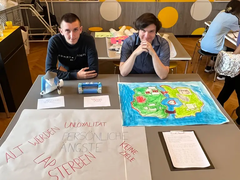 Zwei junge Männer sitzen an einem Tisch. Vor ihnen liegen große Plakate. Eines der Plakate enthält die Worte