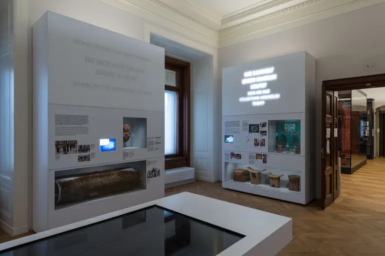 In einem Museum stehen zwei Informationsstände mit Texten und Bildern zu historischen Themen. Einer der Stände hat eine Bildschirmanzeige und ist grafisch gestaltet. Der Boden ist aus Holz, und das Licht fällt durch ein Fenster auf die Exponate.