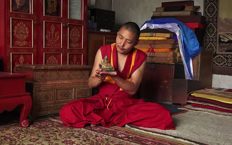 Ein Mönch in traditioneller roter Kleidung sitzt im Schneidersitz und betrachtet eine kleine Buddha-Statue in seinen Händen. Im Hintergrund sind buntgestreifte Kisten und eine verzierte Wand zu sehen. Der Raum hat eine ruhige, meditative Atmosphäre.